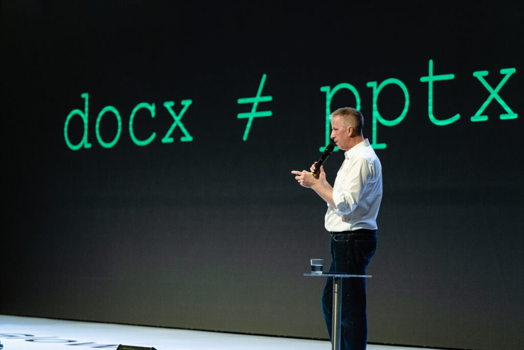 a presenter standing in front of his presentation. the slide describes a rule in presentations: a document is not the same as a presentation. and vice versa